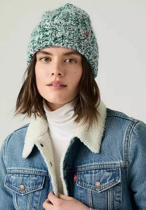 Donna che indossa un berretto lavorato a maglia verde e bianco, un dolcevita bianco e una giacca di jeans blu con colletto in pile bianco su uno sfondo chiaro uniforme.