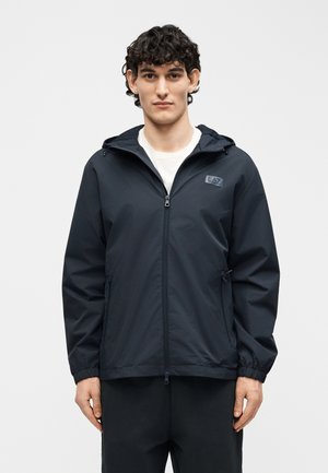 Jeune homme aux cheveux bouclés et foncés, portant une veste à capuche noire zippée sur une chemise blanche, se tenant devant un fond gris clair uni.