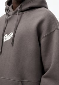 Gros plan d'une personne portant un sweat à capuche gris avec des cordons de serrage et un texte stylisé blanc sur la poitrine.