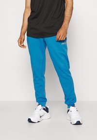 Muž v jasně modrých teplácích ellesse, černém tričku a bílých teniskách Nike, stojící na jednoduchém světlém pozadí.