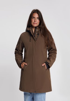 Vrouw met lang bruin haar, gekleed in een bruine capuchonjas met handen in de zakken en lichtblauwe spijkerbroek tegen een effen achtergrond.