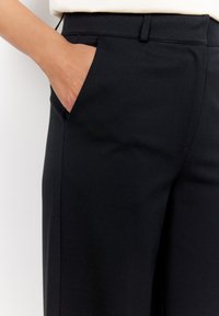Pantalon noir avec une texture lisse, doté d'une poche latérale et d'une coupe ajustée. La taille comprend des passants de ceinture pour une fonctionnalité supplémentaire.