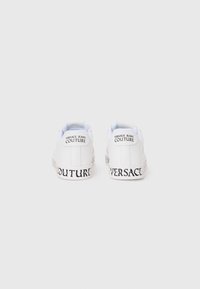 Baltos odinės sportinės bateliai su juodu "VERSACE JEANS COUTURE" prekės ženklu ant nugaros ir pado, pasižymintys lygiu paviršiumi ir apvalia forma.