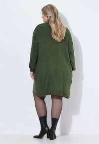 Olivgrünes Strickkleid mit langen Ärmeln, gerippten Bündchen und lockerer Passform. Trägt schwarze Knöchelstiefel und durchsichtige schwarze Strumpfhosen.