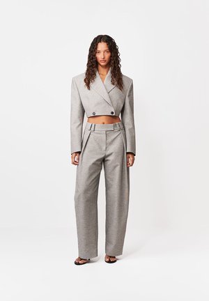 Kvinde med krøllet hår iført en kort, grå, dobbeltradet blazer og højtaljede, vidde bukser, stående på en hvid baggrund.