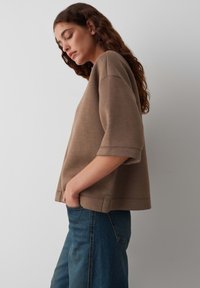 Haut surdimensionné en mélange de coton marron avec des manches courtes, des poches latérales et une coupe décontractée. Associé à un pantalon en denim bleu.