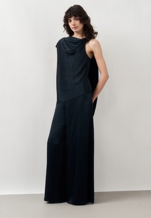 Göndör sötét hajú nő, aki ujjatlan fekete, redős, bőszárú nadrágot visel, egy egyszínű világos fal előtt áll.