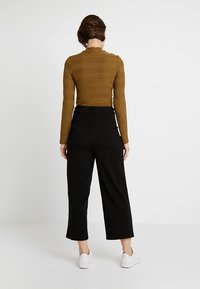 Brun ribbad långärmad topp med hög hals, kombinerad med vida svarta byxor. Modellen bär vita sneakers. Minimalistisk bakgrund.