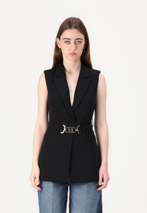 Black sleeveless blazer with a deep V-neck, featuring a gold belt buckle with logo. Smooth texture and tailored design paired with blue jeans.