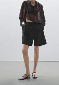 Personne portant un chemisier transparent noir, un short noir taille haute avec les mains dans les poches, un bracelet manchette argenté et des sandales noires avec des détails argentés.