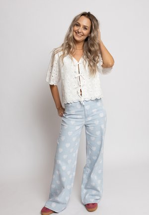 Glunderende vrouw met lang golvend haar draagt een witte blouse met oogjes, lichtblauwe wijde broek met witte hartjes en rode schoenen, poserend tegen een effen achtergrond.