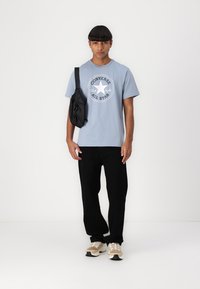T-shirt din bumbac bleu deschis cu logo circular Converse, geantă neagră tip crossbody, pantaloni negri, adidași bej cu accente texturate și șireturi.