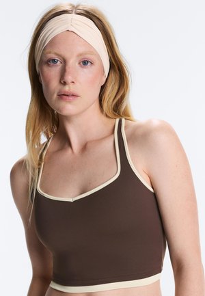 Jonge vrouw met blond haar die een beige hoofdband en een bruin cropped topje met beige rand draagt tegen een witte achtergrond.