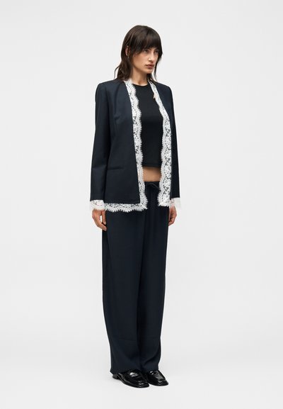 Γυναίκα που φοράει μαύρο παντελόνι με φαρδιά πόδια, μαύρο κοντό τοπ και μαύρο σακάκι με λευκή δαντέλα στα μανίκια και στο μπροστινό μέρος, στέκεται μπροστά σε λευκό φόντο.