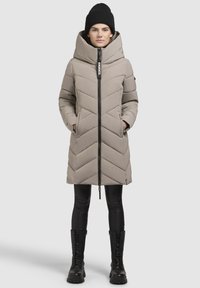 Puffy beige coat with chevron stitching, high collar, and zipper closure. Worn over black leggings and combat boots. Adjustable hood.