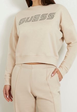 Kvinde iført beige Guess sweatshirt og matchende bukser, stående med den ene hånd i lommen foran en hvid baggrund.
