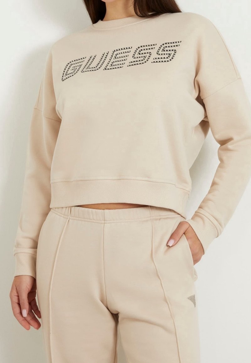 Femme portant un sweat-shirt beige Guess et un pantalon assorti, debout avec une main dans la poche devant un fond blanc.