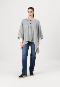 Poncho de lã cinza com um decote redondo, três botões pretos e costura de contraste branca ao longo das bordas. Usado sobre uma camisa branca e jeans.