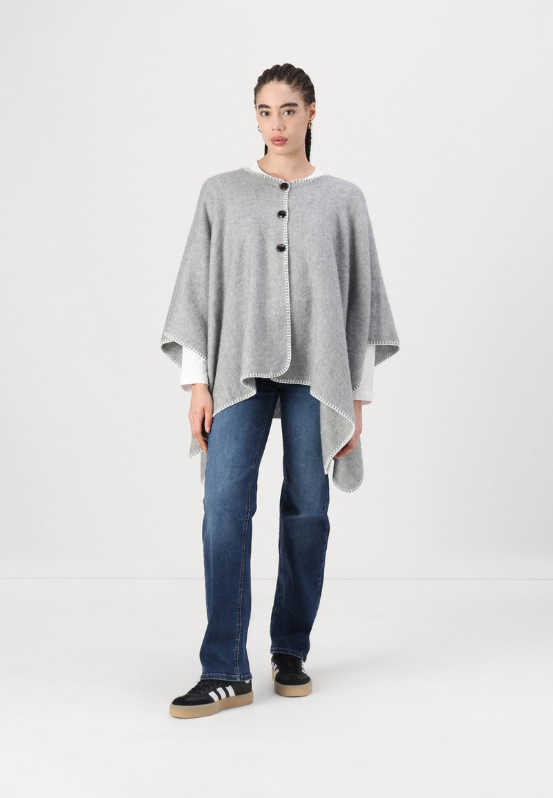 Poncho de lã cinza com um decote redondo, três botões pretos e costura de contraste branca ao longo das bordas. Usado sobre uma camisa branca e jeans.
