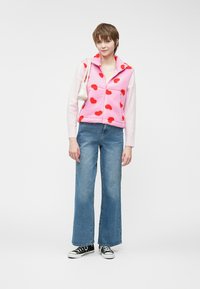 Ροζ φλις γιλέκο με κόκκινα σχέδια καρδιάς, συνδυασμένο με ανοιχτό μπλε φαρδιές τζιν και μαύρα αθλητικά παπούτσια, κρατώντας μία μικρή λευκή τσάντα.