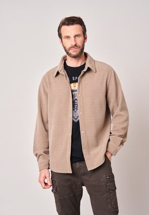 Man met baard die een beige corduroy blouse met knopen draagt over een zwart grafisch T-shirt en donkere cargobroek, staand voor een effen achtergrond.
