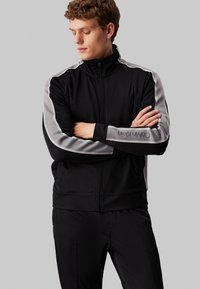 Giacca nera con zip, dettagli grigi ai lati e colletto alto. Realizzata in materiale liscio, presenta una vestibilità slim e un logo sul maniche.