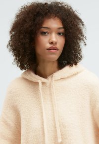 Soft, peach-colored hoodie with a fluffy texture, drawstring detail, and a relaxed fit. Features a hood and a high neck.