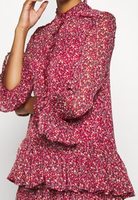 Red floral blouse with a high neck, sheer texture, ruffled cuffs, and a layered hem. Features small multicolored floral patterns.