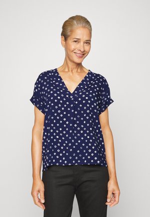 Blusa azul-marinho de manga curta com decote em V, apresentando um padrão geométrico branco. Tecido suave com um corte descontraído, combinada com calças pretas.