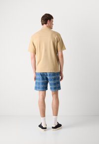 Beige színű, rövid ujjú póló, kék kockás farmer rövidnadrággal párosítva. Mindkét ruhadarab laza szabású. Fekete cipő és fehér zokni egészítik ki a megjelenést.