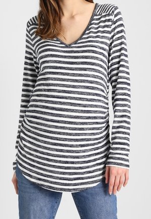 Femme portant un haut à manches longues rayé horizontalement noir et blanc à col en V, associé à un jean bleu, debout sur un fond uni.