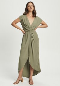 Olive green maxi dress with cap sleeves, deep V-neck, twisted front detail, and asymmetrical hem. Smooth texture. Worn with brown heels.