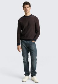Donkere gebreide trui met ribbelstructuur en contrasterende marineblauwe halslijn, gecombineerd met blauwe denim jeans en witte sneakers.