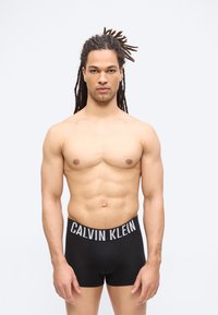 Νεαρός άνδρας με μακριά dreadlocks, φορώντας μαύρα εσώρουχα Calvin Klein boxer briefs, στέκεται μπροστά σε απλό ανοιχτόφωτο φόντο.