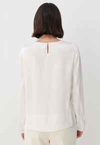 Blusa bianca in satin a maniche lunghe con dettaglio a chiave sul retro, texture liscia e orlo dritto, abbinata a pantaloni rigati color crema.