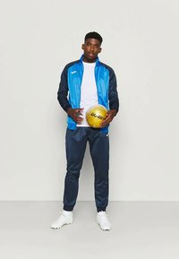 Sportinis apranga, kurią sudaro mėlynas švarkas su juodais akcentais, balta marškinėliai, tamsiai mėlynos treniruočių kelnės ir baltos sportinės bateliai, laikantis auksinį kamuolį.