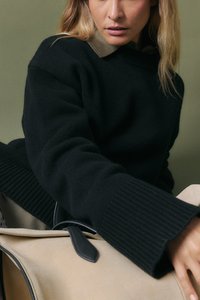 Zwarte gebreide trui met geribbelde manchetten, gedragen over een overhemd met kraag, gecombineerd met een crèmekleurige tas met zwarte accenten.