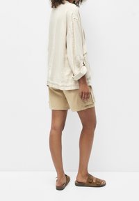Veste oversized beige clair avec des manches retroussées, associée à un short cargo kaki et des sandales marron, sur fond blanc uni.