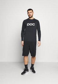 Svart långärmad tröja med "poc"-logotyp, ihop med svarta shorts och svarta träningsskor. Enkelt, slätt tyg och avslappnad passform.