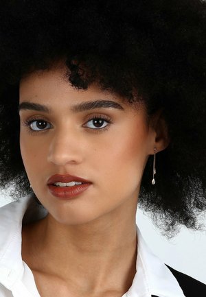 Hängende Ohrringe mit einem birnenförmigen Charm, ausgestattet mit einem metallischen Finish. Das Model hat dunkles, lockiges Haar und ein Make-up in neutralen Tönen.