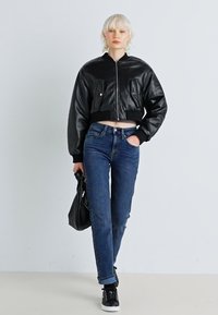 Black leather cropped bomber jacket with ribbed cuffs, two front pockets, paired with blue jeans and black sneakers, holding a black bag.