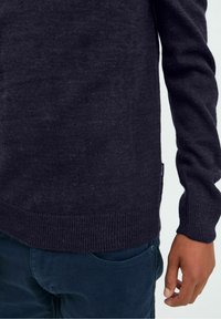 Person in einem dunkelgrauen Strickpullover und dunkelblauen Hosen, die rechte Seite von der Mitte des Oberkörpers bis zur Hand vor einem schlichten Hintergrund zeigend.