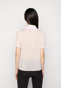 Blouse légère et transparente de couleur blanche avec un motif floral orange, manches courtes bouffantes, et un col en vichy rose froncé. Texture douce et design léger.