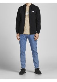 Uomo che indossa una felpa nera con zip, una t-shirt grafica beige, jeans azzurri chiari e sneakers nere, in piedi contro uno sfondo grigio uniforme.