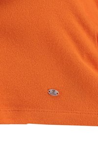 Oranges Strickstoff-T-Shirt mit einem kleinen Metalletikett, auf dem "KEY LARGO" eingraviert ist, glatte Textur und ein einfaches, unaufdringliches Design.