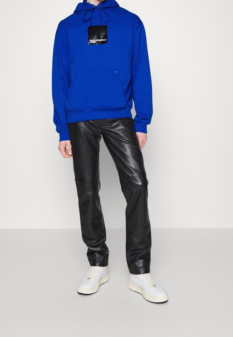 Sudadera azul con cordón y bolsillo frontal; pantalones de cuero negros; zapatillas deportivas blancas; etiqueta visible en la sudadera.