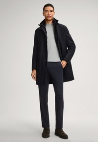 Cappotto navy con colletto alto e chiusura a bottoni, maglione grigio sotto e pantaloni scuri. Il modello si presenta con sicurezza, indossando stivali alla caviglia marroni.
