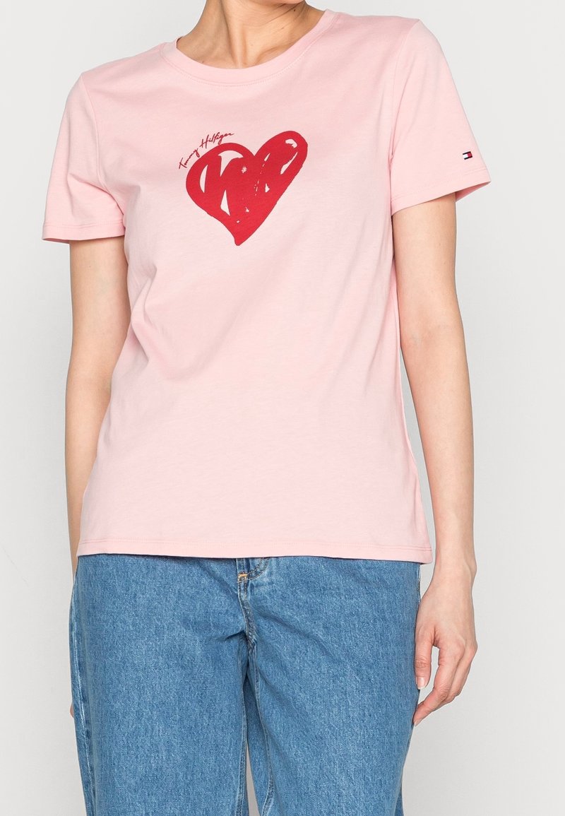 T-shirt en coton rose avec un graphisme de cœur rouge et un design de texte. Manches courtes et col rond. Porté avec un pantalon en denim bleu.