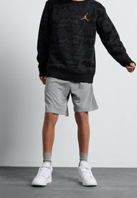 En person står klädd i en svart sweatshirt med ett guldigt logotyp, grå shorts och vita sneakers, mot en ljusgrå vägg och golv.