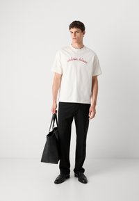 White cotton t-shirt with red "calvin klein" logo, paired with black trousers and a black leather tote bag. Smooth texture, relaxed fit.